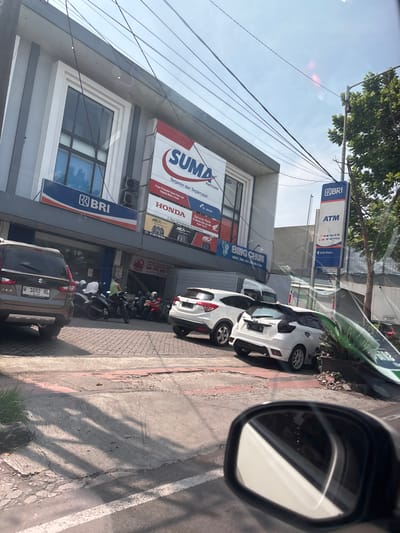The image shows a street scene in Sidoarjo, Indonesia (latitude: -7.442, longitude: 112.687). A gray brick building with a large sign advertising "SUMA Parts Shop" is visible. The sign is red with white text, and there are two smaller signs for HONDA and BRI underneath it. There is a white Honda HR-V car parked in front of the building, and another white car, possibly a Toyota Yaris, is parked behind it. There are several motorcycles parked in front of the building, including some with people sitting on them. The street is made of gray concrete and has a white line painted down the center. There is a large tree on the right side of the street, and a sign for BRI ATM is visible in the image. There are also a few people walking on the sidewalk. The sky is a light blue with some clouds. The scene is shot from the perspective of a car traveling down the road. The car's mirror is visible in the lower right corner of the image.