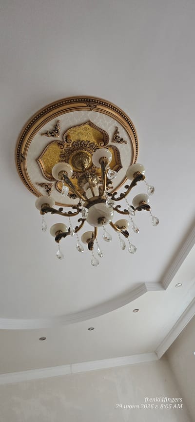 This is a low-angle, indoor, medium shot of a chandelier hanging from a white ceiling with decorative molding. The chandelier is ornate, made of gold-colored metal, and features multiple frosted glass light shades and crystal teardrop accents. The ceiling has a circular decorative medallion behind the chandelier, with gold detailing. The lower portion of the image shows a white wall, possibly with a textured finish, and recessed lighting in the ceiling. The timestamp in the bottom right corner reads "29 июня 2026 г. 8:05 AM" and a username "frenki4fingers," indicating the image was taken in Verkhniy Naur, Russia.