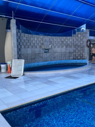 A top-down shot shows a swimming pool and its surroundings. The pool's edge is tiled in white, leading to a blue-tiled pool surface. A small, curved, blue-tiled section parallels the pool. Next to the pool is a tiled walkway leading to a blue structure. The walkway features various tile patterns. A lifeguard float, some construction materials, and a grate are near the walkway. A person is relaxing on a lounge chair in the distance.
