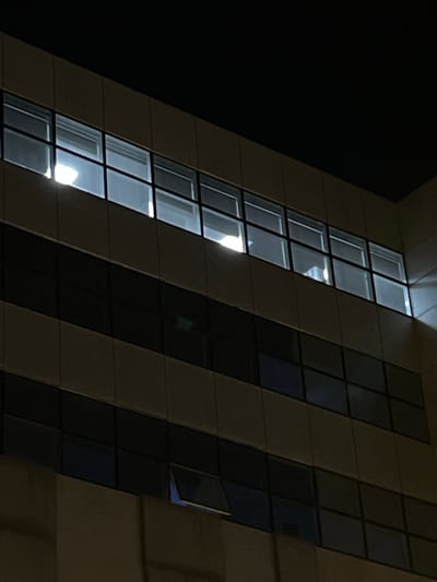 A nighttime view of a building with a modern architectural design. The building features a facade with a pattern of alternating light and dark sections. The light sections are comprised of a series of windows that are brightly illuminated, suggesting activity inside the building. The dark sections appear to be columns or sections of the building's exterior that are not illuminated. One window is slightly ajar. The building is located in Meknes, Morocco.