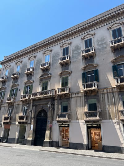 This media file shows the facade of an old building in Catania, Italy. The building has multiple stories with numerous windows, many of which are adorned with small balconies and decorative elements. Some windows have green shutters, while others are open, revealing the interior or reflecting the blue sky. The architecture appears historic, with ornate cornices and detailing along the facade.
On the ground floor, there are storefronts. One storefront clearly displays the name "miramira" in large, illuminated letters, with a display of what appear to be cosmetic or perfume products inside. Another storefront below it also has "miramira" branding, suggesting a retail establishment. Further down, there's another storefront with a sign that partially reads "FREE".
The image is taken from a slightly angled perspective, looking upwards at the building. The bright blue sky suggests it is daytime, and the strong shadows cast by the architectural features and balconies indicate a sunny day, likely mid-morning or early afternoon. The overall scene depicts a street view in a European city, characterized by its classical architecture and commercial activity. There are no people visible in the image. A parking sign and a numbered address sign (111) are visible near the large dark entrance doors.