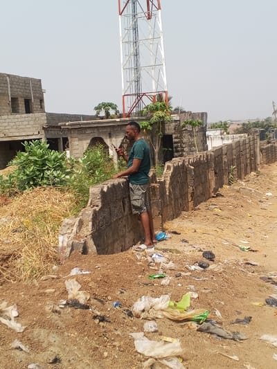 This image captures a daytime scene in Zango-ndaji, Nigeria. A male individual, appearing to be an adult, stands on a dirt path next to a partially dilapidated concrete block wall. He wears a dark green short-sleeved t-shirt, light-colored cargo shorts, and white slide sandals. The man holds a dark-colored smartphone in his right hand, actively looking at its screen, while his left forearm rests on the top of the concrete wall.

The immediate foreground consists of bare reddish-brown earth heavily strewn with litter, including numerous plastic bags, wrappers, and other discarded debris. Behind the man and the wall, dense green shrubs and some dry, yellowish vegetation are visible. In the background to the left, an unfinished two-story building constructed from concrete blocks stands with exposed rebar. To the right of this building, another structure with an arched opening and more vegetation is present. Dominating the upper background is a tall, red and white lattice telecommunication tower against a clear, pale blue sky. Further in the distance, additional buildings and sparse greenery are visible under the bright sky.