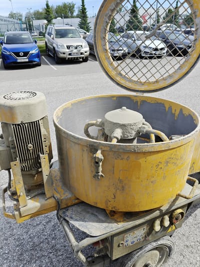 The image shows a yellow concrete mixer parked on an asphalt surface in Tänassilma, Estonia. The mixer is positioned in the foreground, with its electric motor and mixing drum prominently visible. The drum is open at the top, revealing a mixing mechanism covered in dried concrete. The exterior of the mixer is dusty and stained with concrete residue. In the background, several cars are parked in a lot, including a blue SUV and a silver SUV. The area appears to be an industrial or commercial zone, with a building and trees visible in the distance. The sky is overcast with some blue patches. There are no people visible in the image.