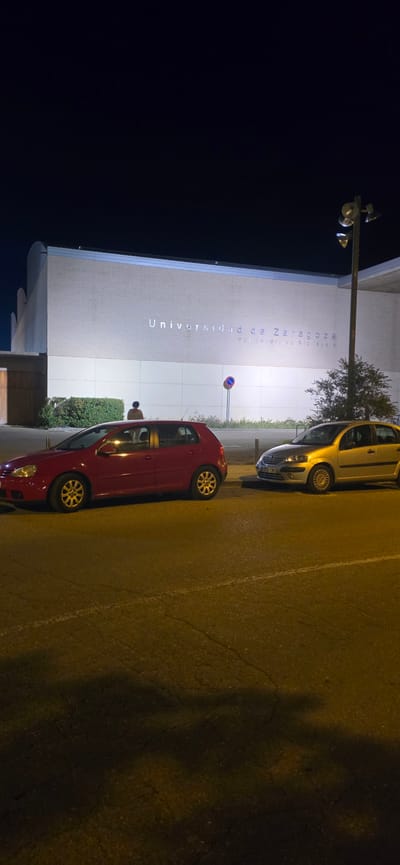 The image depicts the exterior of the "Universidad de Zaragoza" building in Huesca, Spain, at night. The building has a light-colored brick facade with prominent silver lettering spelling out "Universidad de Zaragoza". Below this, smaller text is visible but difficult to read. A single person, seen from the back, is standing near some bushes in front of the building. Two cars are parked on the asphalt road in the foreground: a red hatchback on the left and a silver hatchback on the right. A lamppost with multiple lights is visible on the right side of the image, illuminating the scene. The overall environment is dark, characteristic of nighttime, with artificial lighting from the building and the lamppost.