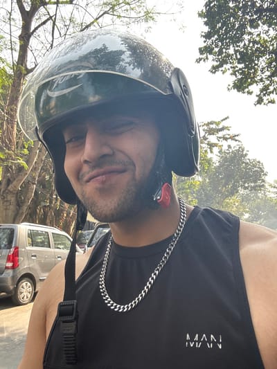 A man wearing a helmet and a black tank top with the word "MAN" visible. He's also wearing a silver chain around his neck. He appears to be outdoors, possibly riding a motorcycle or scooter, with trees and cars in the background. The photo is taken at an angle, showing the man from a low perspective.
