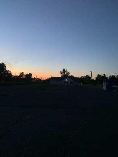 The image depicts an outdoor scene at dusk, likely in Hoholeve, Ukraine. The sky transitions from a deep blue overhead to a soft orange glow near the horizon, indicating the sun has recently set. In the middle ground, a low-slung building with a light on is visible, surrounded by trees and utility poles. To the right, a small, blue and white structure, possibly a kiosk or shelter, stands near a fence. The foreground is dominated by a dark, paved surface, appearing to be a parking lot or open area with visible cracks. The overall atmosphere is quiet and serene.