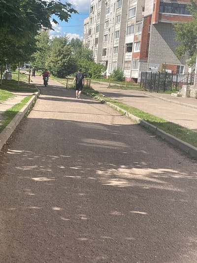 The image shows a paved pedestrian path in a city. On the path, a person wearing a black t-shirt and shorts with a black backpack is walking away from the camera. Further ahead, another person is riding a scooter, also moving away from the camera. The path is lined with grass and a curb, and shadows from trees are cast on the pavement. In the background, there is a multi-story apartment building with windows and balconies, and more trees. This scene is consistent with a residential area in Yaroslavl, Russia, likely during daytime given the bright lighting and shadows.