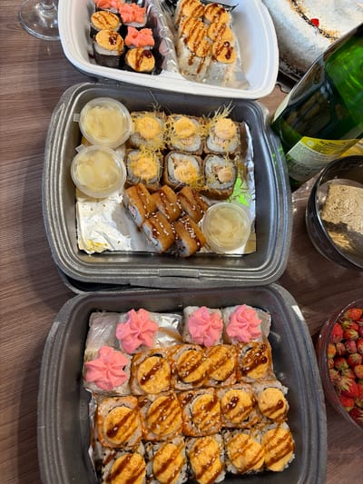 An overhead shot displays a wooden table laden with various food items and drinks. In the upper left, a wine glass is visible. Next to it, a white take-out container holds an assortment of sushi rolls, some topped with pink, decorative cream and others with a browned, creamy sauce drizzled with dark sauce. To the right of this container, another white container features more sushi rolls, predominantly topped with the same browned, creamy sauce. A green bottle of what appears to be sparkling wine stands prominently in the upper right quadrant of the image. Behind the bottle, a white cake decorated with shredded coconut and a red cherry on top is partially visible. Below the first two sushi containers, a dark grey take-out tray contains a variety of sushi rolls. Some are topped with a browned, creamy sauce and sprinkled with shredded cheese or similar toppings, while others are deep-fried and coated in sesame seeds, with a drizzle of dark sauce. Two small plastic containers filled with a light yellow, chopped condiment are placed on top of the sushi in this tray. To the right of this tray, a dark bowl holds a light brown, paste-like substance. In the lower right, a clear glass bowl is filled with small, red wild strawberries. The scene is well-lit, and the focus is sharp, highlighting the textures and details of the food. The image does not provide any specific information about the location within the city of Субханкулово, Russia.