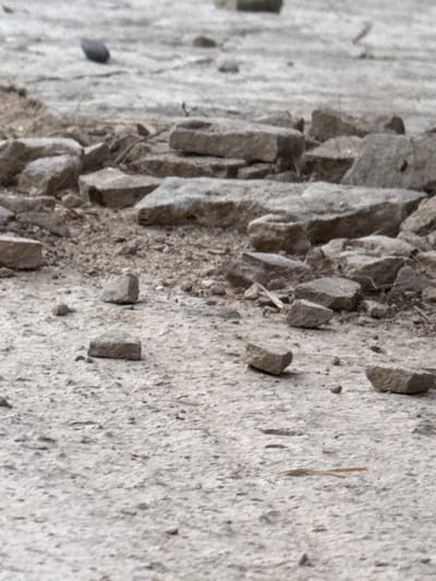 The image shows a close-up, low-angle, slightly blurred view of a dry, rocky, and sandy ground surface. Scattered across the ground are numerous flat, light-colored stones of various sizes, some of which appear to be broken or chipped. The overall impression is one of an arid, undeveloped area. There are no people, animals, or clear man-made structures visible. The scene is devoid of any vegetation, except for possibly a single thin, light-brown twig or reed-like object in the lower right quadrant of the image. The background is a continuation of the same rocky and sandy texture, with more stones and debris. The lighting suggests an overcast or diffused light source, as there are no strong shadows. The ground appears to be a mix of fine gravel and larger stones, with a dusty or dry appearance. There is no indication of water, though the general appearance could be that of a dry riverbed or a barren landscape. The location is not identifiable from the image.