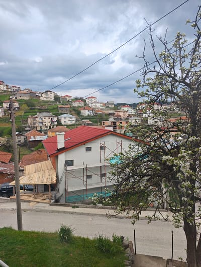 The image shows a hillside residential area in Kasak, Bulgaria, under a cloudy sky. A paved road runs along the bottom of the frame, with grass on either side. A blossoming tree occupies the right side of the image. In the center, a partially constructed two-story building with a red roof has scaffolding erected against its side. To the left, a partially completed wooden structure is visible. In the background, the hillside is dotted with houses of varying sizes and styles, mostly with red roofs. Power lines stretch across the sky above the houses. A utility pole is visible on the left edge of the image. A black car is parked near the wooden structure.