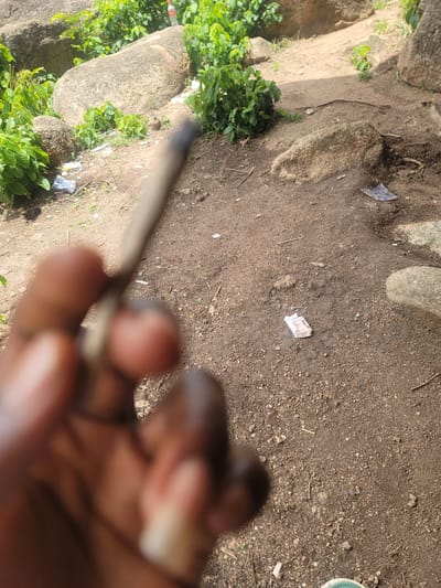 The image shows an outstretched hand holding a rolled cigarette. The background consists of a dirt ground littered with stones, debris, and foliage. The location is Barakin Akawo, Nigeria.