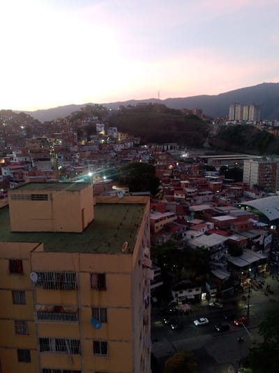 The image shows a dusk aerial view of a densely populated urban area in Caracas, Venezuela, likely a hillside barrio or informal settlement. The foreground features the corner of a light yellow, multi-story apartment building with visible windows, some barred. A green roof with small white objects and a red circular sign is also prominent. Beyond this building, a sprawling cityscape unfolds, characterized by numerous smaller, densely packed residential structures with red and white roofs, many appearing to be built on a steep incline. Lights are visible within many of these buildings, indicating evening activity. Further in the distance, larger modern apartment buildings and some industrial-looking structures are visible, set against rolling hills or mountains that form the city's backdrop. The sky is a pale gradient of light purple and pink, suggesting the sun has recently set. The overall impression is one of a vibrant, albeit informal, urban environment at twilight.