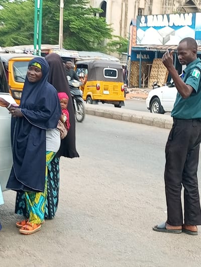 The image captures a street scene in Arawa, Nigeria. In the foreground, a woman wearing a dark blue hijab and a vibrant, patterned skirt is standing, holding a small child in a pink garment. She appears to be looking towards the left, possibly at someone or something off-camera. To the right of the woman, a man in a green uniform with a Nigerian flag emblem on his sleeve is visible. He is looking to his right and holding up his hand, appearing to gesture or communicate. His dark pants and open-toed sandals are also visible.
Behind the individuals, several vehicles are present. A yellow auto-rickshaw, commonly known as a "Keke Napep," is prominently parked on the left. Further back, another similar vehicle and a white sedan are visible. The background features buildings, one of which has a large blue and white sign that reads "HARA FOAM" and also "HARA." There are also trees with green foliage, suggesting it might be daytime with moderate to warm weather. The overall atmosphere suggests a busy street or market area, with people going about their daily activities. The people are dressed in a way that is common for the region.