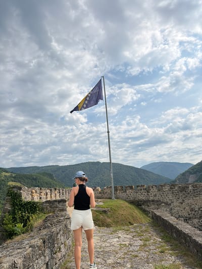 A young woman stands on a stone wall, looking out at a mountainous landscape. She is wearing a black tank top, white shorts, and a blue baseball cap. A Bosnian flag flies on a flagpole to her left. The sky is cloudy, and the sun is shining. The scene appears to be set during the daytime. The woman's posture and gaze suggest she is contemplative or enjoying the view. The stone wall and the flag indicate a historical or cultural site, possibly a fortress or castle, given the location context of Jajce, Bosnia and Herzegovina, known for its historical fortifications. The greenery on the mountainside suggests a temperate climate.