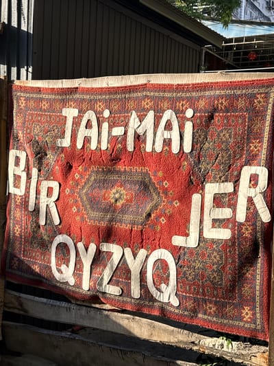The image depicts a red Persian rug hanging outdoors, possibly as a banner or decoration. White letters are attached to the rug, spelling out "TAI-MAI B/R QYZYQ JER". The rug is patterned with traditional geometric designs in red, blue, and yellow. It is attached to a wooden structure, and in the background, a dark, corrugated metal wall is visible, suggesting an urban or industrial setting. The overall impression is that of an outdoor establishment, possibly a cafe, shop, or event space, in Bishkek City, Kyrgyzstan. The lighting suggests it is daytime, with shadows cast by the sun. There are no people visible in the image.