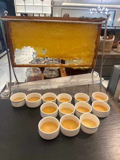 The image shows a honeycomb in a metal frame next to a collection of small white cups filled with honey. The scene appears to be a food display, likely a breakfast or brunch buffet. The setting is presumed to be in Doha, Qatar, based on the visual elements and likely context, though specific evidence within the image to confirm this is not available. Details such as the interior architecture, lighting, and general ambiance suggest a modern, upscale establishment. No people are visible in the image.