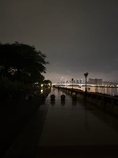 The image depicts a waterfront promenade at night in Tianhe District, China. The promenade is wet, suggesting recent rain, and is illuminated by streetlights and the city lights across the water. On the left side of the promenade, silhouetted trees and foliage line a grassy embankment. Several spherical decorative objects are placed along the edge of the promenade. Further along the promenade, a few people are visible walking. Across the water, a cityscape is illuminated, with buildings and a bridge lit up. The sky is dark and overcast.