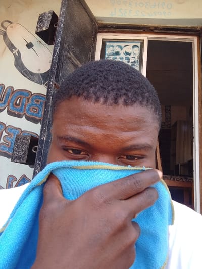A dark-skinned individual is depicted in a close-up, with their face partially obscured by a light blue microfiber towel held by their right hand, covering the nose and mouth. The individual's short, dark, curly hair and eyes, directed downwards, are visible. They wear a white shirt or t-shirt. The scene is located in Zango-ndaji, Nigeria, outside what appears to be a barbershop. To the left of the subject, a light-colored exterior wall displays a large painted image of a barber's clipper in white and black, with partial blue block lettering visible below it. A dark, possibly metal, utility box is mounted on the wall above the painting. To the right, an open doorway or window with a white frame reveals the interior of a building. Inside, a poster showcasing various men's hairstyles is affixed to the wall. Above the doorway, faint, reversed, or stylized script is present. Deeper within the interior, shelving units and a light-colored curtain are visible.