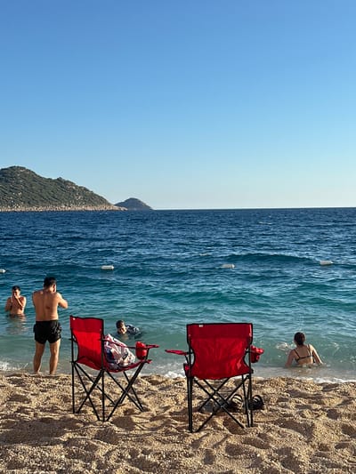 The image captures a vibrant, sunny day at a pebbly beach. The scene is divided horizontally into three main sections: a clear blue sky at the top, a broad expanse of sparkling blue sea in the middle, and a light-colored pebbly beach at the bottom. In the distance on the left horizon, a verdant, mountainous landmass is visible under the clear sky.

Several people are enjoying the water. On the right side of the frame, a woman in a dark bikini is partially submerged at the water's edge, seemingly wading or entering the sea. Towards the left, two men are standing in the shallow, clear water. One man, shirtless and wearing dark swim shorts, has his back to the viewer. Another person is partially submerged next to him. Further towards the center-right, near a red chair, a person or child is also in the water, their head visible above the surface.

Two red folding beach chairs are set up on the pebbly shore, close to the water. One chair on the right has a pair of dark sandals placed beside it. Another chair towards the center has a bag or towel resting on it, with some red fabric (possibly from another bag or item) nearby. The water is a brilliant turquoise close to the shore, transitioning to a deeper blue further out. Several white buoys are scattered across the water's surface in the middle distance, indicating a designated swimming area or boat lane.

The weather is bright and clear, suggesting midday or early afternoon. The overall impression is one of leisure and enjoyment of a beautiful, clear-water beach. No discernible text is visible on any objects.