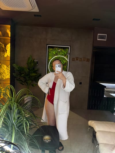 A woman, seen in a mirror reflection, stands in a spa or wellness center, holding a smartphone to take a selfie. She is wearing a red one-piece swimsuit and a white waffle-knit bathrobe. Her hair is pulled back, and she has a necklace on. She is positioned in front of a wall with a green, vertical garden display and a light fixture. To her left, a large potted plant is visible, and further left, a display case with yellow decorative items. To her right, a dark wooden structure, possibly part of a sauna or changing room, and a cushioned lounge chair are in view. The floor is tiled with large, light-colored tiles. The overall ambiance suggests a tranquil and modern spa environment. The image was taken in Chingeldi, Uzbekistan.