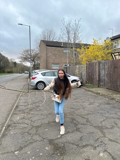 The image depicts an outdoor scene on an overcast day in Corby, United Kingdom. A female subject runs or strides towards the camera on an asphalt surface. She has long, dark brown hair and is dressed in a light-colored fuzzy jacket over a dark top, light blue skinny jeans, and light-colored sneakers. Her mouth is open, and her arms are outstretched. In her left hand, she holds a long, slender, multi-branched wooden stick. Her right hand grips a white disposable cup with a dark lid. A small, light-colored shoulder bag with a distinct emblem is worn across her body.
The asphalt ground displays numerous cracks and signs of wear. To the left of the subject, a curb delineates the pavement from a road. Several silver hatchback cars are parked along the road. A grey street lamp post stands on the left. In the background, a two-story brick residential building with a dark roof and a satellite dish is visible behind a dark brown wooden fence. Bare deciduous trees are present to the left and behind the building, while a tree or bush with bright yellow blossoms is visible behind the fence to the right. The sky is uniformly overcast.