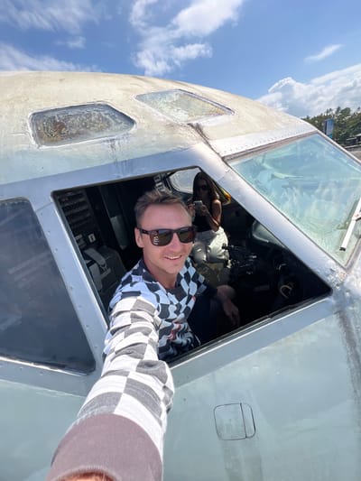 In Blahbatuh, Indonesia, a man with sunglasses and a checkered long-sleeved shirt is taking a selfie from the cockpit of a derelict airplane. He is smiling and positioned in the pilot's seat, extending his arm to capture the shot. Behind him, a woman wearing sunglasses and a black tank top is seated in the co-pilot's seat, looking at her phone. The cockpit interior is visible, showing various controls and gauges. The airplane's exterior appears weathered and worn, with a cloudy blue sky and lush green vegetation visible in the background.