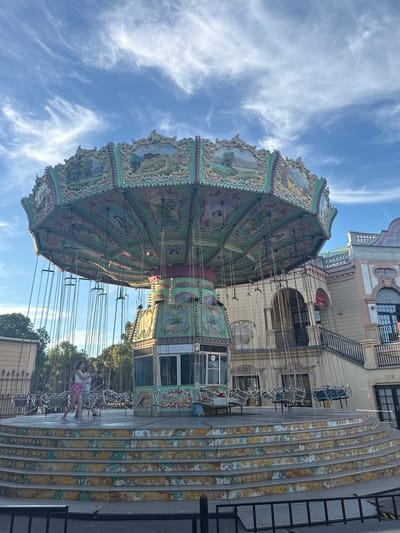 This media file captures a low-angle view of a carousel swing ride on a bright, cloudy day. The ride is elaborately decorated with ornate green and gold trim, featuring painted panels with picturesque scenes. The carousel's structure is visible, with numerous chains hanging down, some supporting empty seats. In the background, a multi-story building with architectural details and windows can be seen.
Two young girls, dressed in summer clothing, are standing on the carousel's platform. One girl wears a white top and a pink patterned skirt, while the other is in a white top and pink shorts. They appear to be waiting or have just exited the ride.
The sky is a vibrant blue, filled with wispy white clouds, suggesting fair weather and daytime. The scene is set at what appears to be an amusement park or fairground, given the presence of the carousel ride and the surrounding buildings which might house other attractions or shops. The overall mood seems to be cheerful and summery, evoking a sense of fun and leisure. The context of Vienna, Austria, implies a European setting for this amusement. No visible text is present in the image.