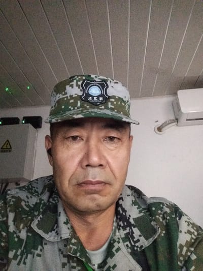 A close-up, chest-up, front-facing photograph features a middle-aged East Asian man wearing a camouflage military uniform and a matching cap. The cap displays a circular emblem with text and a graphic that appears to be a parachute. The man has short, dark hair, brown eyes, and is wearing a black earpiece in his left ear. The background shows a textured ceiling and a white wall with an air conditioning unit. To the left of the man, a gray electrical box with a yellow warning symbol is visible, along with two green indicator lights. The lighting is moderate, and the image has a slightly grainy quality. The scene is indoors, suggesting an office or technical room environment. The provided location "Түндүк айланып өтүүчү жол 84 га, Kyrgyzstan" is not directly depicted in the visual elements of the image.
