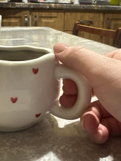 A close-up photograph captures a light-skinned human hand, exhibiting short, clear-polished fingernails, firmly gripping the handle of a white ceramic creamer or small pitcher. The vessel is adorned with multiple small, red heart-shaped decals. This scene is situated on a table covered by a light-toned, possibly off-white, patterned tablecloth. The background, out of focus, reveals wooden kitchen cabinetry with visible handles and a portion of a dark-stained wooden chair, indicative of a domestic kitchen or dining environment in Overyata, Russia. The action depicted is the manual grasping of the creamer.
