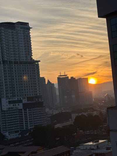 The image captures a sunset over Kuala Lumpur, Malaysia. In the foreground, a tall, modern apartment building with a dark facade and numerous windows dominates the left side of the frame. The building features a helipad at its summit, and visible signage indicates it is "HILTON RESORT SUITES."
Further into the city, a cluster of high-rise buildings forms the urban skyline. Among them, a partially constructed building with exposed scaffolding stands out. The sun, a bright orb, is setting behind these buildings and a distant mountain range, casting a warm, orange glow across the sky and illuminating the clouds with streaks of yellow and orange. The sky exhibits a layered, wispy cloud pattern, typical of an approaching sunset.
The lower portion of the image reveals a denser urban landscape with smaller buildings, some with red tiled roofs, and a dense arrangement of trees casting dark shadows. The overall impression is an urban panorama at dusk, with the natural beauty of the sunset contrasting with the man-made structures of the city. The lighting suggests it is late afternoon or early evening. There are no people visible in the image.