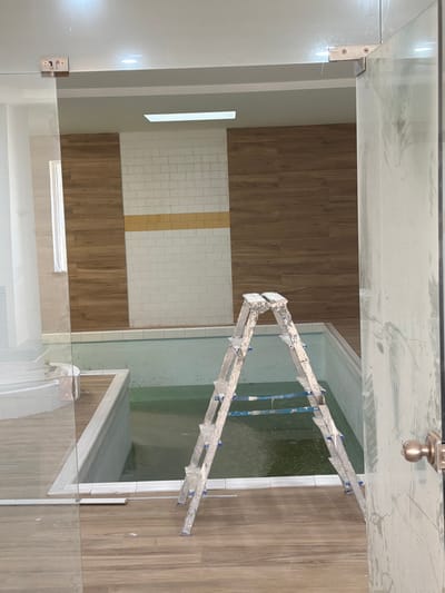 The image depicts an indoor swimming pool area under construction, likely a mikvah, situated in Gondomar, Portugal. A silver aluminum stepladder, showing signs of paint splatters, stands open and centered within the pool's enclosure. The pool itself is filled with greenish water and appears to have a concrete basin. The surrounding walls are finished with a combination of wood-effect paneling and white subway tiles, accented by a horizontal band of yellow tiles. The floor is covered in wood-look laminate flooring. A glass partition with metal hinges is visible on the left, and a doorframe with a brass doorknob is on the right, suggesting a completed section of the building. The ceiling features recessed lighting. There are no people present in the image.