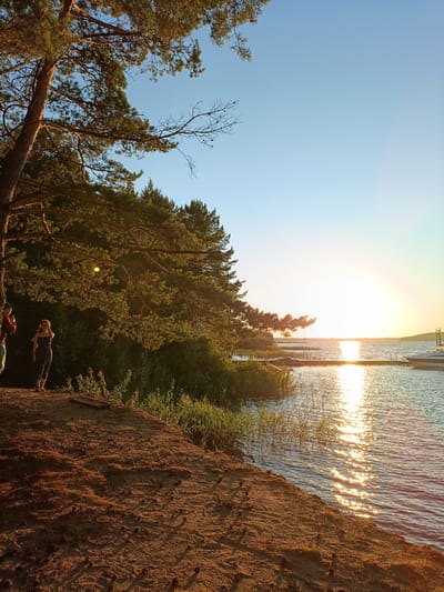 The image depicts a scenic sunset view on the Kama River in Chaykovsky, Russia. In the foreground, a sandy embankment is covered with pine cones and sparse dry grass. Two individuals are visible on the embankment; one person, wearing a dark patterned dress, stands facing the water, while another person, partially obscured, is to their left. Lush green vegetation and pine trees line the embankment, with sunlight filtering through the branches. The river stretches out to the horizon, reflecting the golden hues of the setting sun, creating a shimmering effect on the water's surface. A wooden dock with a boat is situated on the right side of the river. The sky is a clear, pale blue, transitioning to a brighter yellow near the horizon where the sun is setting.