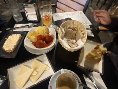 The image depicts a table at a bar or restaurant, possibly in Jerez de la Frontera, Spain, based on the "Cruzcampo" beer brand and typical Spanish tapas. A person's hand is visible on the right. The table is laden with various dishes: a piece of bread with a white spread, a plate of meatballs in sauce and potato chips, a basket of bread, a plate of sliced cheese, and a piece of fried food, perhaps a tortilla. A cup of coffee or tea is also present, along with silverware. Other objects include a water bottle, a napkin dispenser, and a smartphone. A menu or paper is also visible. The environment is a restaurant or bar setting.