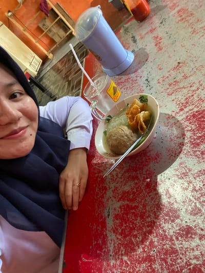 A selfie is taken by a woman wearing a hijab and a white shirt, seated at a red, worn table. She is positioned on the left side of the frame, looking towards the camera with a slight smile. In front of her, on the table, is a bowl of bakso (Indonesian meatball soup) and a glass of iced drink with a straw. A blue dispenser is also visible on the table. The background shows an orange wall and wooden shelves with items, suggesting an indoor dining setting, potentially a local eatery in Sungai Sipai, Indonesia. The lighting appears to be artificial, casting shadows on the table.