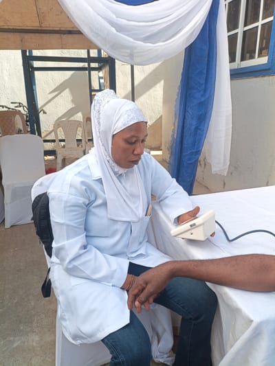 A woman wearing a white lab coat and a white headscarf is seated at a table covered with a white cloth. She is holding a white medical device connected by a black cord to a person's arm. The arm is dark-skinned and clad in blue jeans. The woman is looking down at the medical device. In the background, there are white-covered chairs, white and blue draped fabric, and a building with a window. The location is Tarauni, Nigeria.