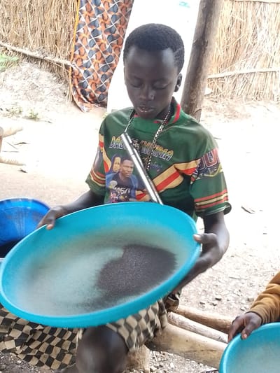 A young boy in Otulu, Nigeria, is shown holding a large, light blue basin filled with dark granules, possibly for mining or processing. He wears a green t-shirt with an image of two men and some text. The t-shirt also features colorful striped patterns on the sleeves and neckline. A silver chain with a pendant hangs around his neck. He is wearing a checkered skirt or wrap.
The background suggests an outdoor setting, possibly a rural or informal settlement. A wall constructed of dried reeds or straw is visible behind him, along with wooden poles supporting some structure. A patterned fabric, possibly a curtain or textile, hangs on the left side of the frame, featuring geometric shapes in orange, blue, and black. The ground appears to be dirt and gravel.
To the boy's left, a blue basin is partially visible, and to his right, another person's hands are seen reaching for a similar blue basin, indicating others are participating in or observing the activity. The overall lighting suggests it is daytime, but the weather is not discernible. The boy's gaze is directed downwards at the basin, suggesting concentration on the task at hand. The image captures a moment of work or a common activity in the depicted environment.