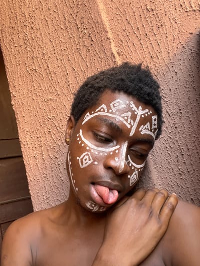 The image features a dark-skinned person with a shaved head lying on a textured, brown surface. White tribal-like designs are painted on their face. The person's eyes are closed, and their tongue is sticking out. An earring is visible on their left ear. The background suggests an outdoor setting, potentially a wall or ledge. The location is Nkpor, Nigeria, based on the user's prompt.