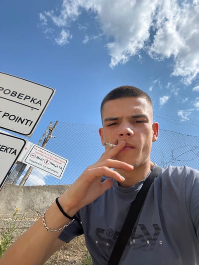 The image is a selfie of a young man with a short haircut, holding a lit cigarette to his mouth. He is wearing a grey t-shirt and a black strap across his chest. He has a bracelet on his left wrist. In the background, there is a chain-link fence with barbed wire, a concrete wall, and several signs. One sign is in Bulgarian and indicates that "On the premises, a permit regime is being enforced! Vehicles and individuals are subject to inspection." Another sign has the word "CHECKPOINT!" on it. The sky is blue with some clouds. The overall setting appears to be an outdoor checkpoint or border crossing, potentially in or near the city of Sopot, Bulgaria, given the Bulgarian text on the sign.