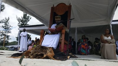 The scene takes place in Rukoko, Democratic Republic of the Congo. A man is seated on a throne with carved lion heads on the armrests. He is dressed in a white robe and a leopard-print hat. He is reading a book and holding a microphone in his left hand. The throne is decorated with a leopard skin and a large, ornate carved wooden top. There are a number of other people seated in the background, some wearing traditional clothing. Several white tents are erected behind the throne, and many trees are visible in the background. The ground is covered with white sand. The scene appears to be an outdoor event, possibly a royal ceremony.