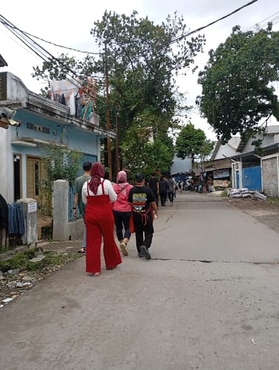 The image depicts a street scene in Katapang, Indonesia, located at the coordinates -6.984 latitude, 107.556 longitude. A group of seven people are walking down a paved street lined with houses and trees. The person in the center of the frame is wearing a black shirt with a yellow logo that reads "SENILAI MERDEKA." The other people are wearing various clothing including red pants, a red shirt, and a blue shirt. The street is bordered by a concrete curb on both sides and there are trees on both sides of the street. The houses in the background are made of concrete and have blue paint. The houses have a variety of architectural styles, including single-story and two-story buildings. There are several electrical wires strung across the street.