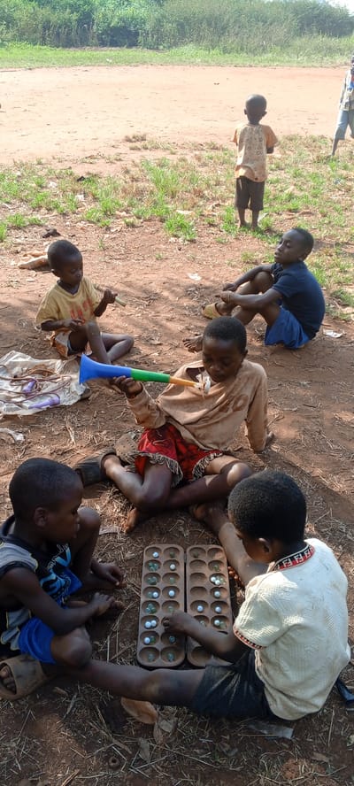 In Ngalula, Democratic Republic of the Congo, six African children are situated on reddish-brown soil scattered with small patches of green grass, twigs, and leaves. The children are positioned in a partial circle. Two are engaged in playing a traditional board game, likely Mancala or a similar variant, on a wooden board with small divots filled with marbles. One child holds a blue and green vuvuzela-like horn. Another child sits nearby with a similar, smaller, unicolored instrument. A length of braided cord is near the vuvuzela. One child is sitting apart, observing the others. Another child is standing alone in the background. The backdrop features sparse shrubbery and a dirt road.