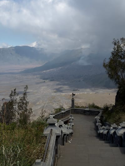 The image depicts a viewpoint overlooking a vast, arid landscape, likely a caldera, with a smoking volcano in the background. In the foreground, a grand stone staircase with ornate balustrades descends into the scene. The staircase is flanked by overgrown, dry vegetation. A single viewing scope is positioned on a stone pedestal to the left of the stairs, suggesting a tourist observation point. The sky is overcast with dramatic clouds. The location is identified as Cemorolawang, Indonesia, a village known for its proximity to Mount Bromo, a famous active volcano. The overall impression is one of natural grandeur and a popular tourist destination for viewing volcanic landscapes.