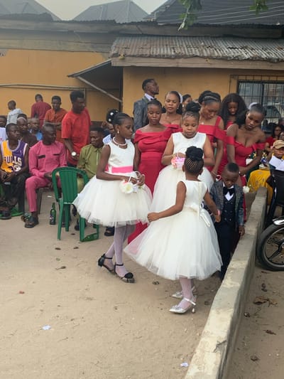 The image captures an outdoor social gathering in Umuode, Nigeria. In the immediate foreground, three young girls in white, layered dresses are visible. The leftmost girl wears a white dress with a pink sash, white patterned tights, and black flat shoes, holding a small white floral basket. The central girl, facing left, wears a white dress with a white sash, white patterned tights, and silver low-heeled shoes, also holding a small white floral basket. She is holding the right hand of a young boy to her right. This boy wears a dark grey sequined suit jacket, a black vest, black trousers, and a black bow tie.
Behind these children, multiple adult women are present, uniformly dressed in off-the-shoulder red formal gowns. Further into the midground and background, numerous men are gathered. Some men are seated on green plastic chairs; one in a pink long-sleeved tunic and matching trousers, and another in an olive-green long-sleeved tunic. Another man in the midground wears a purple and yellow "BULLS 23" basketball jersey. Other men stand or observe in the background, one visible in a red t-shirt and another in a grey patterned suit jacket.
The ground surface is unpaved, consisting of light brown dirt or sand, with scattered leaves and small pieces of debris. Empty green glass bottles are visible on the ground near the plastic chairs. A low concrete curb or barrier runs along the right side of the frame, with the front wheel of a motorcycle partially visible beyond it. The background consists of single-story buildings with mustard-yellow painted walls and weathered corrugated metal roofs. Sparse green foliage is visible overhead in the upper right. The lighting suggests daytime.
