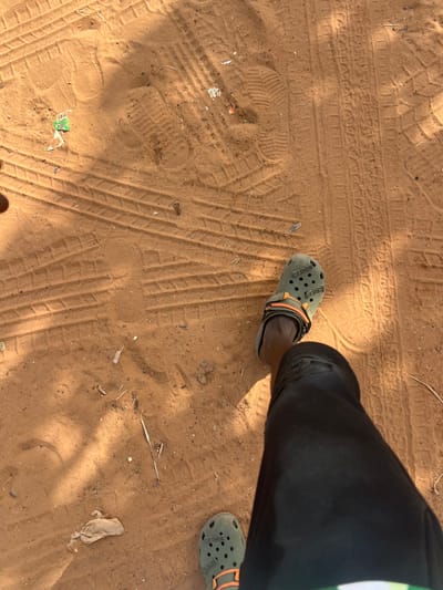 The image displays a ground-level perspective of an unpaved, sandy surface, likely a path, in Birnin Kebbi, Nigeria. A person's lower extremities are visible in the foreground, consisting of dark-colored trousers and two olive green Crocs-brand clogs with orange heel straps. The left foot is partially cropped at the bottom of the frame, while the right foot is more fully depicted. The reddish-brown sand substrate features numerous tire tracks from various vehicles, multiple human footprints (including distinct shoe prints and fainter barefoot impressions), and sparse natural debris consisting of dry plant matter. Scattered anthropogenic litter, including small white and green plastic fragments, is also present. Harsh shadows indicate direct overhead sunlight, characteristic of a daytime outdoor scene.