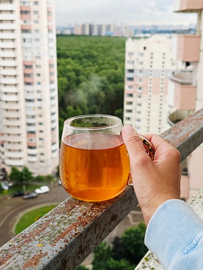 A close-up shot focuses on a hand holding a clear glass mug filled with amber-colored liquid, likely tea, on a rusty metal railing. The mug has a decorative golden rim with star patterns. The background is blurred, showing a city skyline with tall apartment buildings and a dense green forest. The railing appears weathered, with patches of rust and moss. The setting suggests a balcony or elevated outdoor space overlooking a residential area and natural greenery. The weather appears overcast. This scene is located in the city of Odintsovo, Russia.