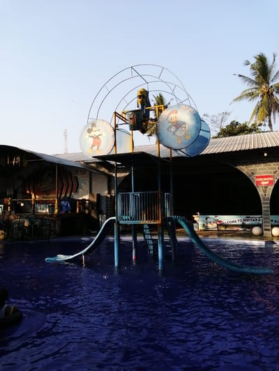 This image depicts a water park slide structure with Mickey Mouse and Donald Duck imagery, located in Kebonsari, Indonesia. The slide, made of blue and green plastic, is positioned over a deep blue pool of water. The structure is supported by blue poles, and a metal fence encloses the top. In the background, a building with a corrugated metal roof houses a stage with "MUSIK ANNIVERSARY" signage. Palm trees and a clear sky are visible above. The scene suggests a recreational environment, possibly during the day, with the pool water appearing calm and reflective.