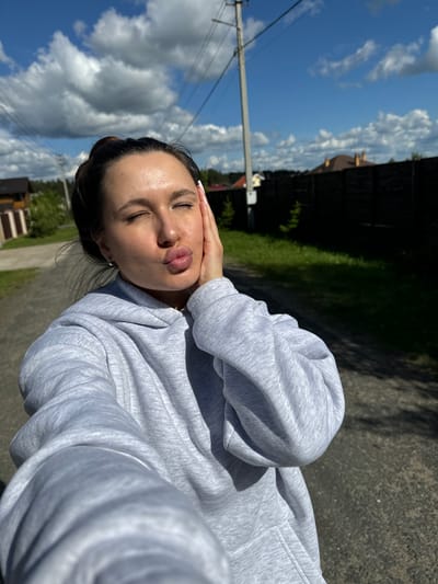 A woman with dark hair pulled back and closed eyes poses outdoors. She wears a gray hooded sweatshirt and has her hand on her forehead. The background shows a road, grass, trees, houses, a power pole, and a blue sky with clouds. The image appears to have been taken on a sunny day. The location may be Vysokinichi, Russia.