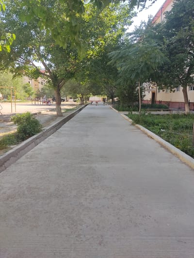 The image shows a concrete walkway in Asmansay, Uzbekistan, lined with trees and bordering a residential building. On the left side of the walkway, there is a children's playground with yellow metal structures. Further down the walkway, several dogs are visible. The residential building appears to be a multi-story apartment complex with visible windows and balconies. Greenery, including bushes and grass, is present along the edges of the walkway and in front of the building. The sky is clear and the scene appears to be set during daylight hours.