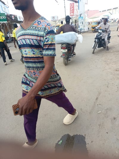 The image depicts an urban street scene in Fagge C, Nigeria, during daylight. In the foreground, partially visible from the chest down on the left, is a dark-skinned male subject walking towards the right. He wears a short-sleeved, V-neck tunic with blue, orange, and white geometric and floral patterns, purple trousers, and beige slip-on shoes. He holds a brown smartphone with a prominent camera module in his right hand. The road surface beneath him is asphalt with patches of dirt, and a wet spot with a green wrapper is visible.
In the midground, a dark motorcycle travels away from the camera, carrying a large white, possibly fabric-wrapped bundle secured to its rear rack; the rider wears a dark shirt. To its right, another dark motorcycle approaches from the background, ridden by a person wearing a light-colored, long-sleeved garment.
The background features multiple commercial buildings. On the left, signage for "OPPO" and "KGS" is visible. On the right, a vertical red sign displays the word "SALE" in white letters, adjacent to a white utility structure, possibly a fuel pump. A yellow auto-rickshaw is discernible further back on the right. Other indistinct human figures are present throughout the background. The sky appears bright and overcast.