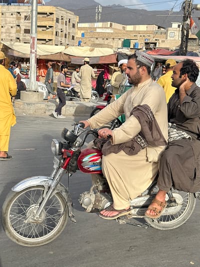 A medium shot depicts two men on a motorcycle in Quetta City Tehsil, Pakistan. The driver, wearing a cream-colored robe and a decorated cap, has a grey scarf draped around his shoulders. He wears brown sandals. The passenger behind him is dressed in brown clothing and brown sandals, with his hand near his face. The motorcycle has a red fuel tank and a silver front fender. In the background, there are buildings, tents, and other people. Some individuals are wearing traditional clothing, and there are flags visible. The overall scene suggests a busy street or market area. Mountains are visible in the distance.